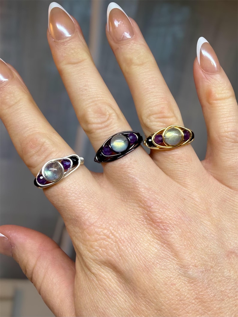 May include: Three wire-wrapped rings with gemstone accents. The rings feature a central, iridescent stone flanked by smaller purple stones. One ring has a silver wire, another black, and the third is gold. The rings are displayed on a hand.