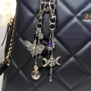 Op de afbeelding: Twee zilverkleurige tashangers hangen aan een zwarte gewatteerde handtas. Eén bedel heeft een vlinder, een facetkristal en zwarte kralen. De andere heeft een halve maan hanger, een pentagram en paarse kralen.