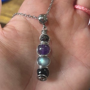 May include: A silver chain necklace with a silver bead and a pendant made of wire wrapped around four gemstones. The gemstones are amethyst, moonstone, black onyx, and hematite.