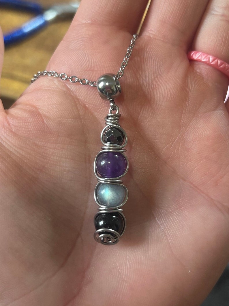 May include: A silver chain necklace with a silver bead and a pendant made of wire wrapped around four gemstones. The gemstones are amethyst, moonstone, black onyx, and hematite.