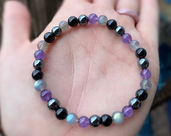 May include: A beaded bracelet featuring black, grey, and purple beads. The bracelet is made from hematite, labradorite, and amethyst.