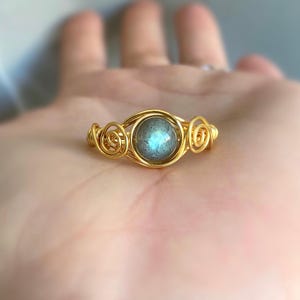 May include: A gold-coloured wire-wrapped ring with a round, iridescent blue-grey gemstone. The ring's design incorporates decorative swirls on either side of the gemstone, creating an elegant and unique piece of jewellery.