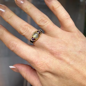 May include: A gold-toned ring featuring a central, oval-shaped, light-colored stone, flanked by two smaller, dark red stones. The ring has a unique design with a black band and is worn on a hand with manicured nails.