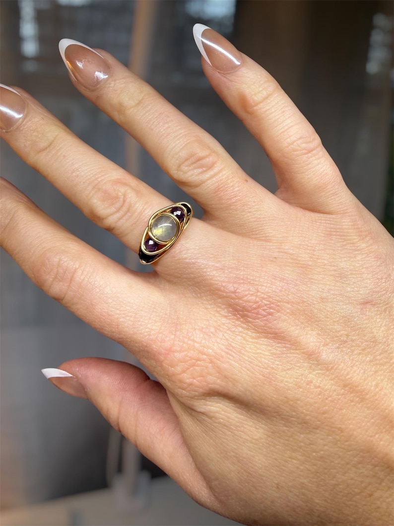 May include: A gold-toned ring featuring a central, oval-shaped, light-colored stone, flanked by two smaller, dark red stones. The ring has a unique design with a black band and is worn on a hand with manicured nails.