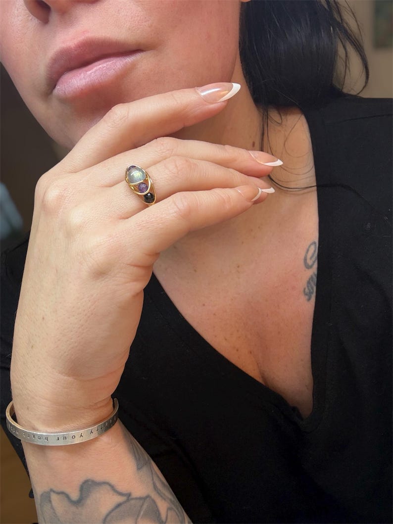 May include: A gold ring with three gemstones: a large oval-shaped light green stone, a smaller purple stone, and a small black stone. A silver bracelet with the text "bury your heart" is also visible.