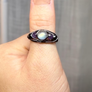 May include: A handcrafted wire-wrapped ring featuring a central, iridescent moonstone flanked by two purple amethyst stones. The ring has a dark, possibly black, wire setting. The ring is on a finger.