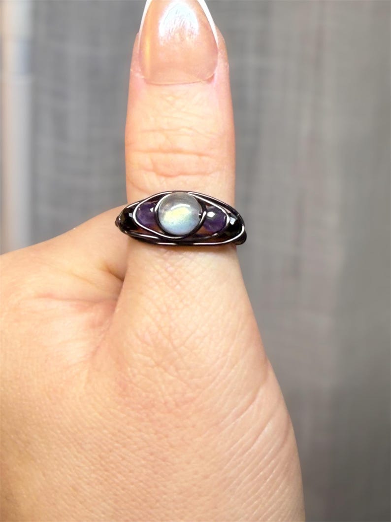 May include: A handcrafted wire-wrapped ring featuring a central, iridescent moonstone flanked by two purple amethyst stones. The ring has a dark, possibly black, wire setting. The ring is on a finger.