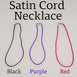Puede incluir: Collares de cordón de satén en negro, morado y rojo. Cada collar tiene un cierre plateado. El texto "Satin Cord Necklace" está en la parte superior de la imagen, con los colores etiquetados debajo.