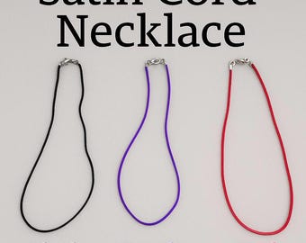 May include: Three satin cord necklaces featuring a black, a purple, and a red cord. Each necklace includes a silver-toned metal lobster claw clasp. The text "Satin Cord Necklace" appears at the top, with "Black", "Purple", and "Red" below.