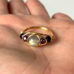 May include: A handmade gold wire ring featuring a central, round, iridescent stone, flanked by two purple and two black gemstone beads. The ring is displayed on a light background.