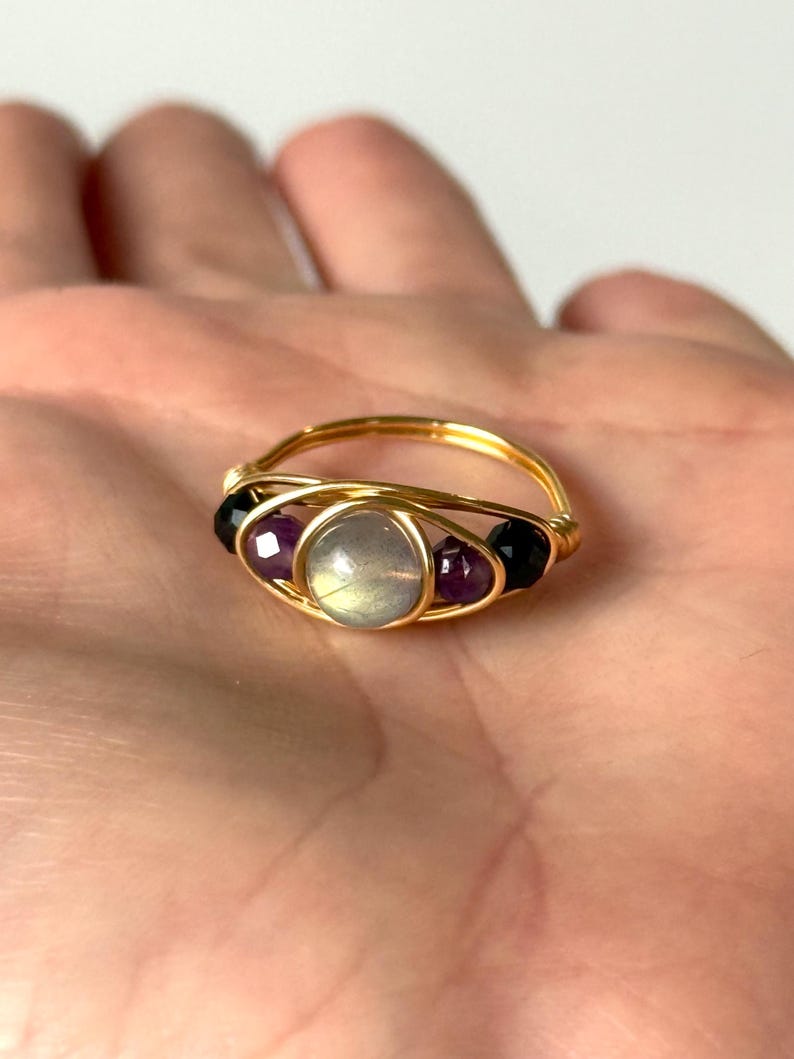 May include: A handmade gold wire ring featuring a central, round, iridescent stone, flanked by two purple and two black gemstone beads. The ring is displayed on a light background.