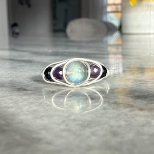 May include: A silver wire-wrapped ring featuring a central iridescent stone, flanked by two purple amethyst stones and two black onyx stones. The ring is set against a blurred background.