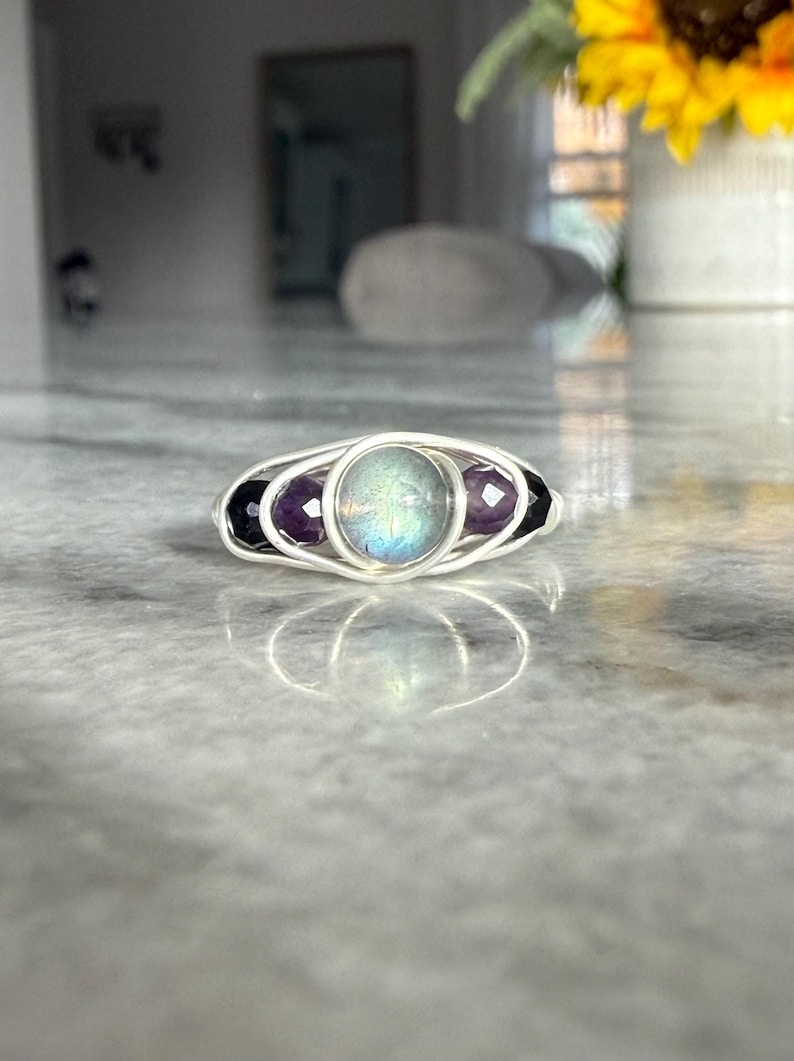 May include: A silver wire-wrapped ring featuring a central iridescent stone, flanked by two purple amethyst stones and two black onyx stones. The ring is set against a blurred background.