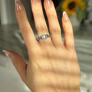 May include: A silver ring featuring a central, round, light-colored stone, flanked by two smaller, oval, dark purple stones. The ring is on a hand with French-manicured nails. The background is blurred, with a glimpse of a sunflower.