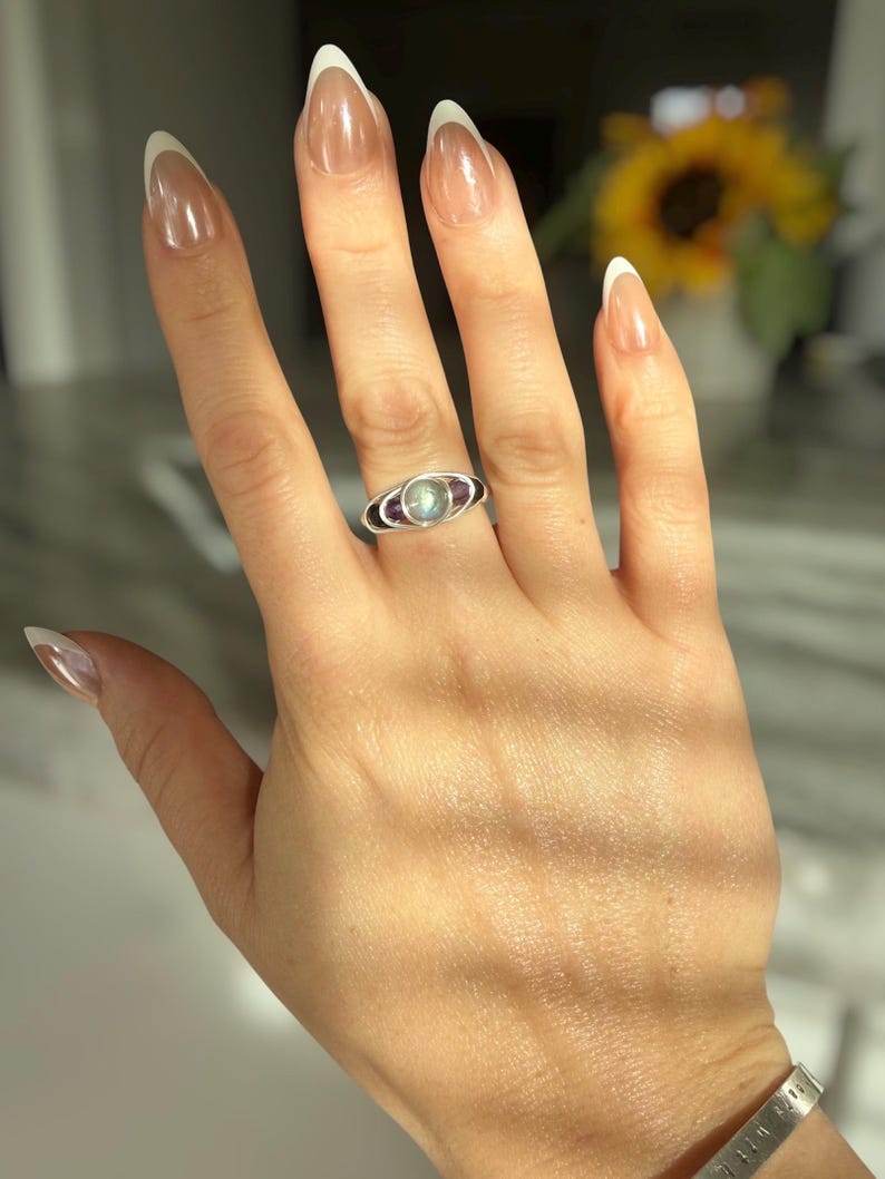 May include: A silver ring featuring a central, round, light-colored stone, flanked by two smaller, oval, dark purple stones. The ring is on a hand with French-manicured nails. The background is blurred, with a glimpse of a sunflower.