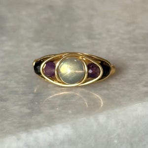 May include: A gold-toned ring featuring a central oval-shaped, iridescent stone, flanked by two smaller, purple gemstones. The ring's design incorporates a wire-wrapped setting, creating a delicate and intricate appearance. The ring is set against a light-colored surface.