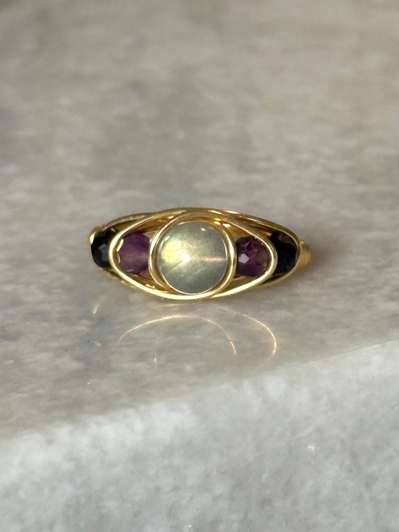 May include: A gold-toned ring featuring a central oval-shaped, iridescent stone, flanked by two smaller, purple gemstones. The ring's design incorporates a wire-wrapped setting, creating a delicate and intricate appearance. The ring is set against a light-colored surface.