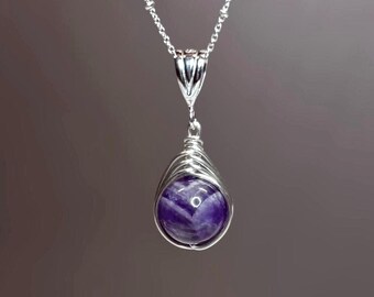 May include: A silver pendant necklace featuring a teardrop-shaped, purple amethyst gemstone wrapped in silver wire. The gemstone is suspended from a delicate silver chain. The necklace is displayed against a neutral background.