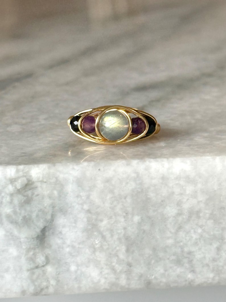 May include: A gold-toned ring featuring a central, round, iridescent stone, flanked by two purple stones and two black stones. The stones are set within a woven gold wire design. The ring is displayed on a light-colored surface.