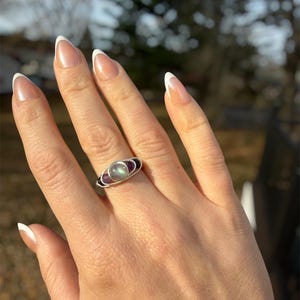 May include: A silver ring featuring a central, oval-shaped, light-colored stone, flanked by two purple stones. The ring is set against a hand with manicured nails. The ring's design includes a dark band on either side of the central stone.