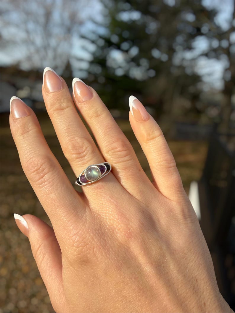 May include: A silver ring featuring a central, oval-shaped, light-colored stone, flanked by two purple stones. The ring is set against a hand with manicured nails. The ring's design includes a dark band on either side of the central stone.