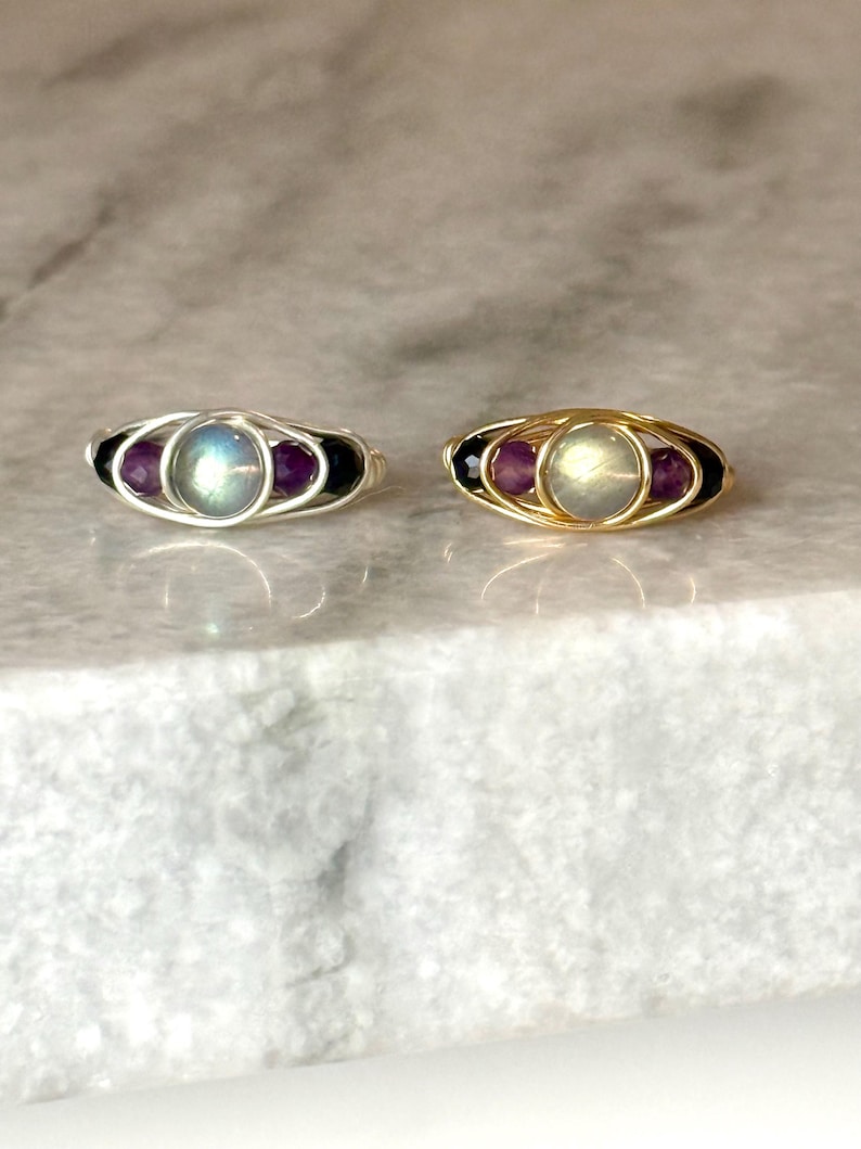 May include: Two wire-wrapped rings, one silver and one gold, each featuring a central iridescent stone flanked by purple and black beads. The rings are displayed on a light-colored, marbled surface, showcasing their intricate design and contrasting colors.