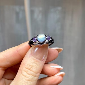 May include: A handcrafted wire-wrapped ring featuring a central, iridescent, oval-shaped stone, flanked by two smaller, purple, round stones. The ring has a dark, possibly black, wire frame. The ring is held up by a person's fingers.