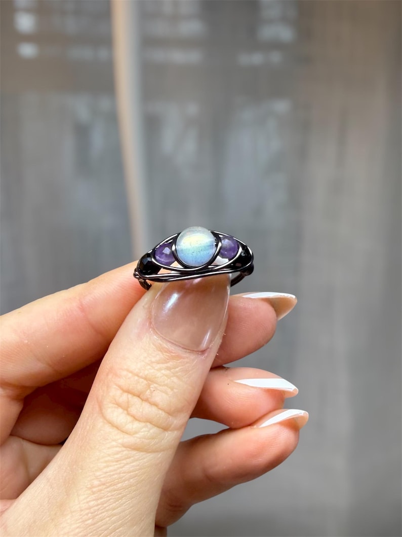 May include: A handcrafted wire-wrapped ring featuring a central, iridescent, oval-shaped stone, flanked by two smaller, purple, round stones. The ring has a dark, possibly black, wire frame. The ring is held up by a person's fingers.