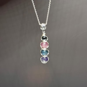 May include: A silver necklace featuring a pendant with a spiral design and a series of colorful gemstones. The gemstones include black, pink, blue, and purple stones. The necklace hangs against a dark gray background.
