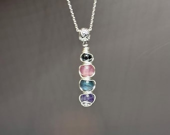 May include: A silver necklace with a pendant composed of a spiral design and a sequence of coloured gemstones. The gemstones are black, pink, blue, and purple. The necklace is set against a dark grey backdrop.