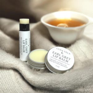 以下が含まれることがあります： Luna VelaのEarl Greyリップバームの画像。リップバームスティックと、開いたものを含む2つの小さなバーム缶が写っています。「Earl Grey Lip Balm」と書かれたラベルには成分が記載されています。背景にはお茶のカップがあります。