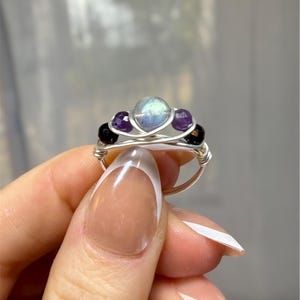 May include: A handmade wire-wrapped ring featuring a central iridescent stone, flanked by two purple amethyst stones and two black onyx beads. The ring has a silver-colored wire band and a delicate, intricate design.