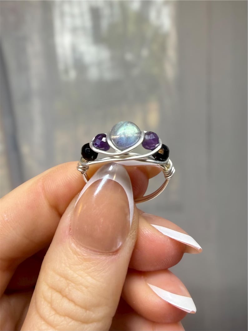 May include: A handmade wire-wrapped ring featuring a central iridescent stone, flanked by two purple amethyst stones and two black onyx beads. The ring has a silver-colored wire band and a delicate, intricate design.