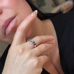 May include: A silver wire-wrapped ring featuring a central, round, iridescent stone, flanked by two purple stones on each side. The ring is displayed on a finger, showcasing its intricate design and craftsmanship. The ring's design is delicate and elegant.
