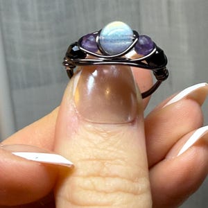 May include: A handcrafted wire-wrapped ring featuring a central light blue stone, flanked by two purple stones and two black beads. The ring's wire is dark brown, creating a contrast with the colorful stones. A unique piece of jewelry.