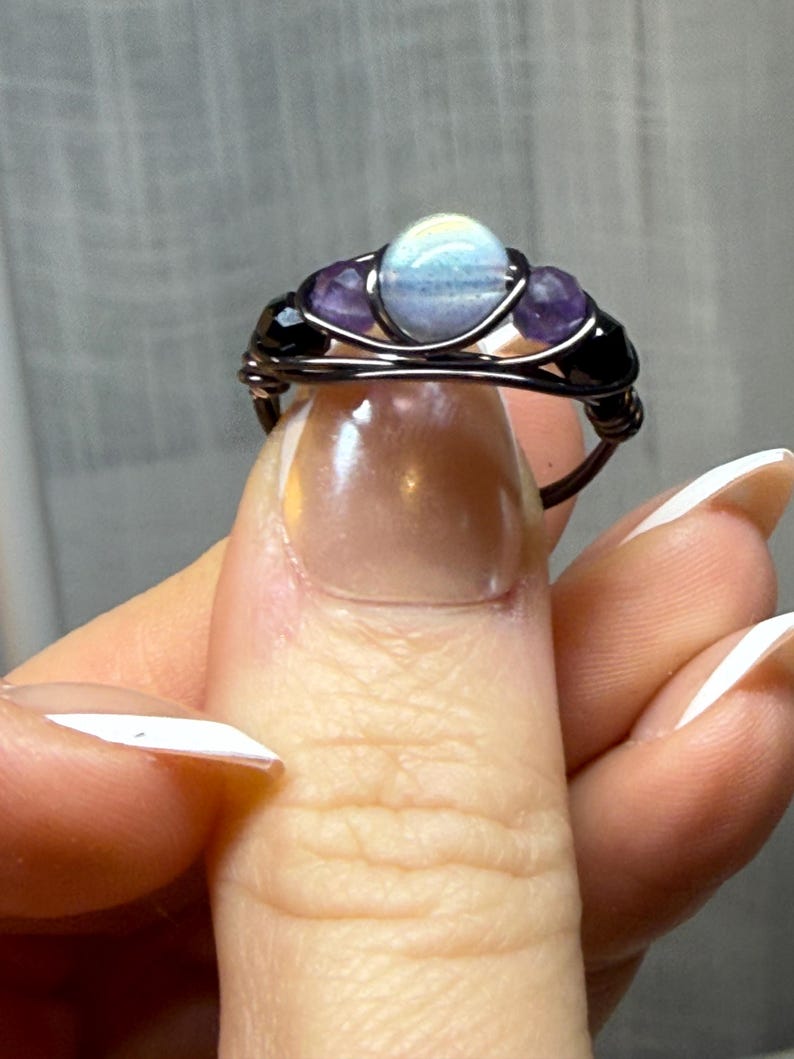 May include: A handcrafted wire-wrapped ring featuring a central light blue stone, flanked by two purple stones and two black beads. The ring's wire is dark brown, creating a contrast with the colorful stones. A unique piece of jewelry.
