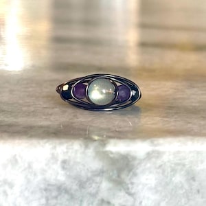 May include: A handcrafted ring with a wire-wrapped design. The ring features a central, round, light green stone, flanked by two smaller, oval, purple stones. The wire is a dark, metallic color, creating a contrast with the colorful stones.