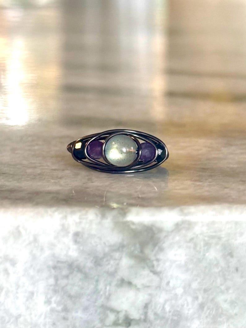May include: A handcrafted ring with a wire-wrapped design. The ring features a central, round, light green stone, flanked by two smaller, oval, purple stones. The wire is a dark, metallic color, creating a contrast with the colorful stones.