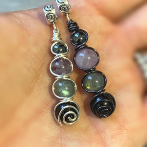 May include: Two silver charm pendants with black and silver wire wrapped around various colored gemstones. The gemstones include amethyst, rose quartz, labradorite, and black onyx.