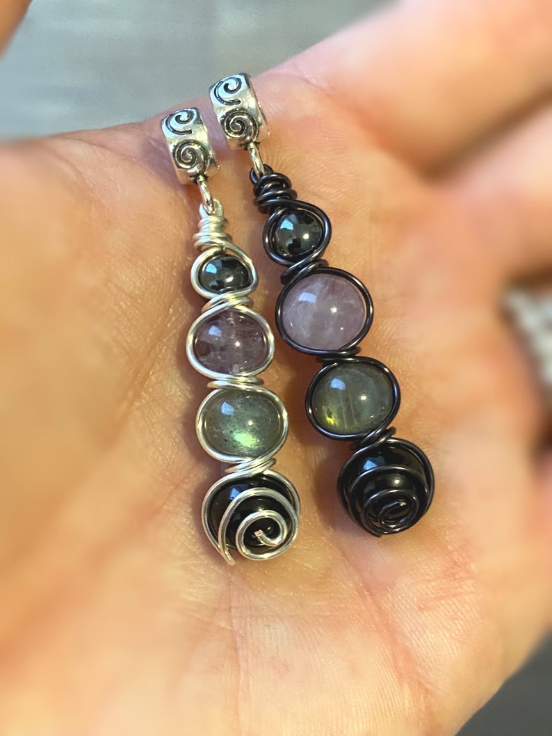 May include: Two silver charm pendants with black and silver wire wrapped around various colored gemstones. The gemstones include amethyst, rose quartz, labradorite, and black onyx.