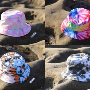 May include: Four different tie-dye bucket hats in pastel, rainbow, space, and black marble patterns. The hats are photographed on a sandy beach.