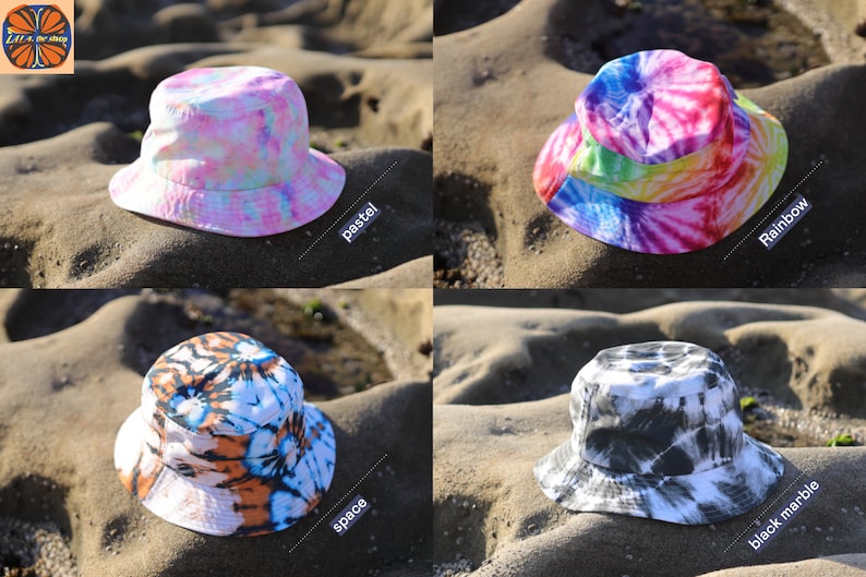 May include: Four different tie-dye bucket hats in pastel, rainbow, space, and black marble patterns. The hats are photographed on a sandy beach.