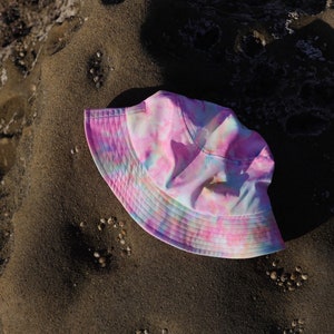 May include: A pink, blue, and white tie-dye bucket hat. The hat is lying on a sandy surface.