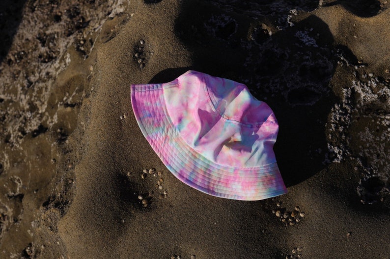 May include: A pink, blue, and white tie-dye bucket hat. The hat is lying on a sandy surface.