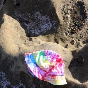 May include: A tie-dye bucket hat with pink, blue, yellow, and green colors. The hat is lying on a sandy surface.