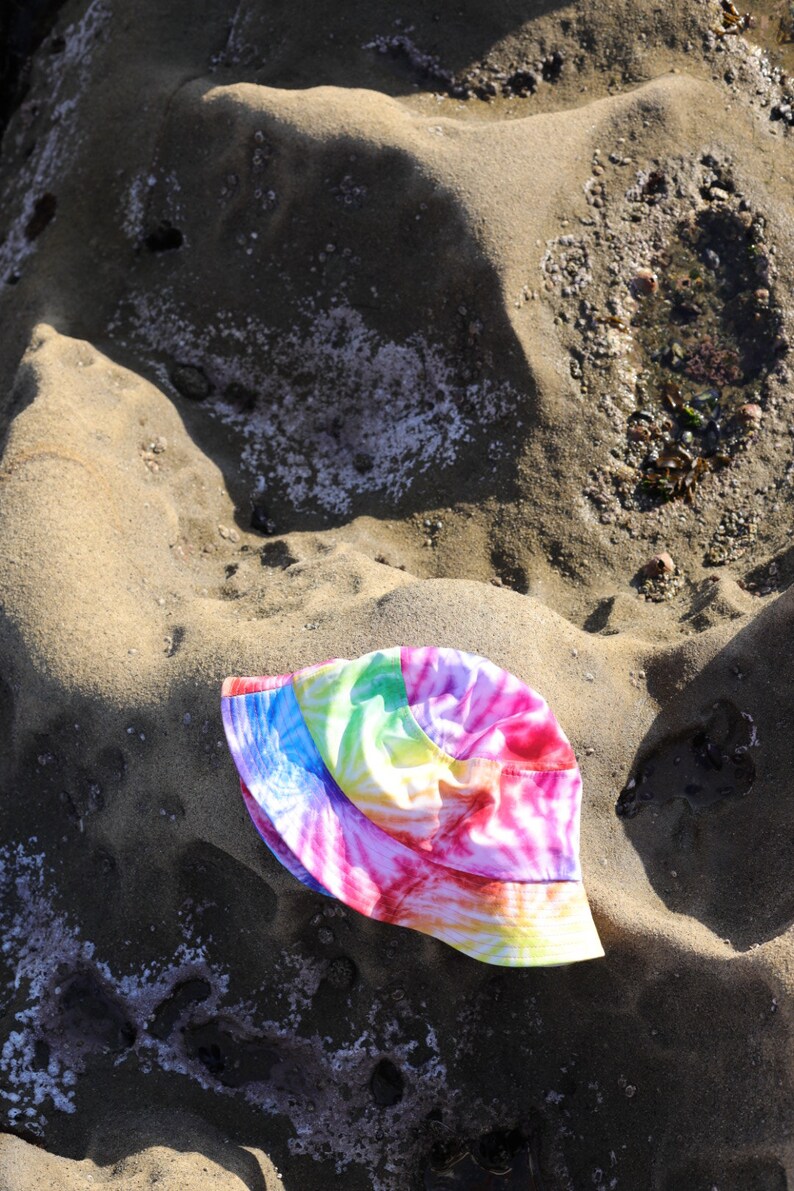 May include: A tie-dye bucket hat with pink, blue, yellow, and green colors. The hat is lying on a sandy surface.