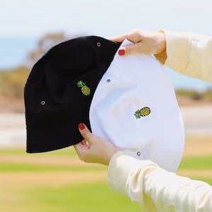 May include: Two bucket hats, one black and one white, with a pineapple embroidered on each. The hats are held by a person's hands.