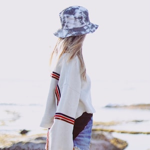 May include: A woman wearing a white sweatshirt with red and blue stripes on the sleeves, denim shorts, and a black and white tie-dye bucket hat. She is standing on a rocky beach.