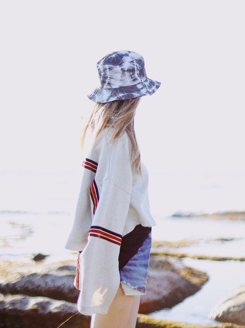 May include: A woman wearing a white sweatshirt with red and blue stripes on the sleeves, denim shorts, and a black and white tie-dye bucket hat. She is standing on a rocky beach.