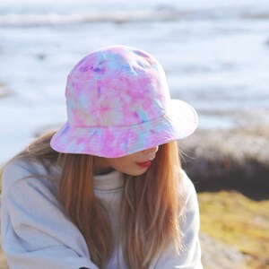 May include: A person wearing a pink, blue, and white tie-dye bucket hat. The hat has a wide brim and is made of a soft, comfortable fabric.
